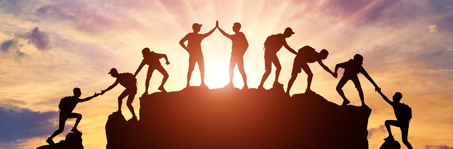 Several people climbing a mountain at sunset. They are helping each other and high-fiving.