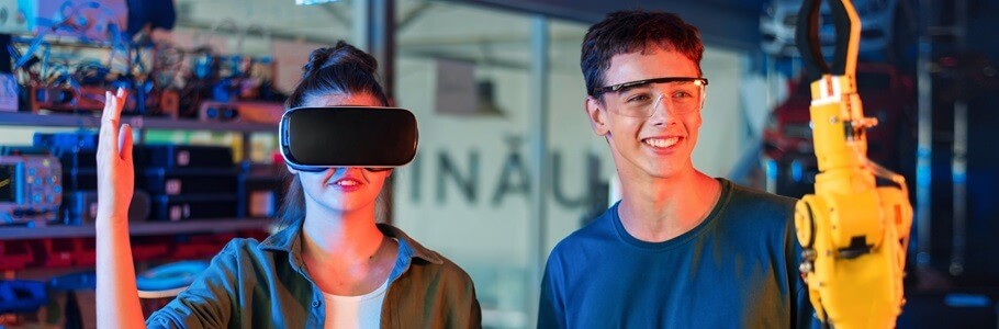 A student wearing a VR headset, alongside a second student looking at a machine arm.